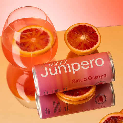 Can of San Junipero Blood Orange soda with a glass of the drink and blood orange slices on a peach background