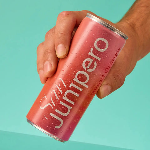 Hand holding a pink can of San Junipero Blood Orange drink against a teal background